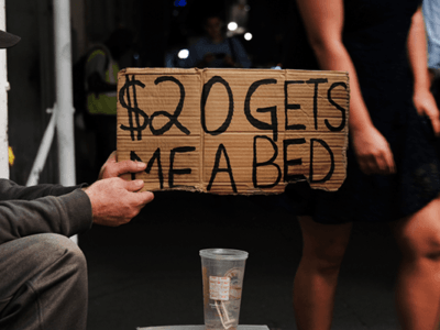 Man holding cardboard sign saying "$20 get me a bed"